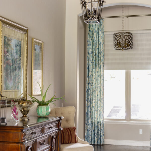 A well-decorated living room with elegant sheer blue floral curtains, high-end roman shades and a sparkling chandelier hanging from the ceiling.