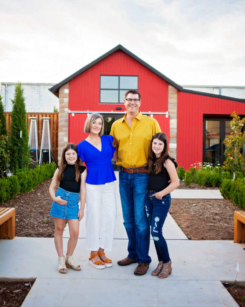 Heather Langhofer with family smiles in front of a red structure, capturing a precious moment. she is the owner of Grace Allen Design, a luxury window coverings business in Edmond, Oklahoma.